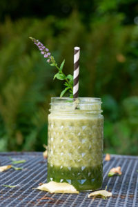 Grüner Smoothie im Glas mit Strohhalm und Minzblüte steht auf Gartentisch, Hintergrund unscharf, grün. Grüner Smoothie im Glas mit Strohhalm und Minzblüte steht auf Gartentisch, Hintergrund unscharf, grün.